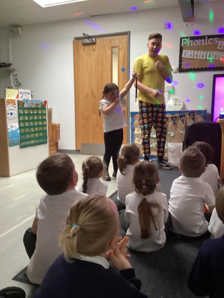 Children sat on the floor watching an entertainer and a child stood at the front of the room together